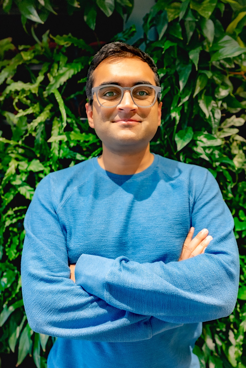 Chandim Chatterjee, wearing glasses and a blue sweater, standing in front of green foliage.